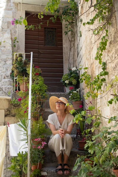 Mooie vrouwelijke toerist draagt grote stro zonnehoed en sundress zitten en ontspannen op oude stenen huis trappen tijdens de zomer reizen op de Middellandse Zee kosten op warme zomerdag. — Stockfoto