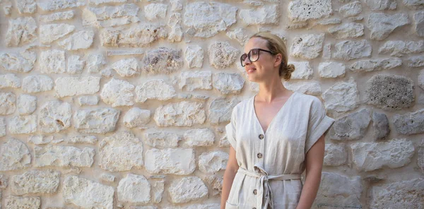 Porträt einer schönen, fröhlichen blonden Frau mit einteiligem Sommerkleid und Sommerhut, die vor einer alten mittelalterlichen Steinmauer steht. Sommerferienkonzept. — Stockfoto