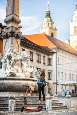 Gezgin, çevre dostu elektrikli scooterla eski kasabayı keşfediyor. Ljubljanas 'ı keşfeden kadın turist eski ortaçağ tarihi şehir merkezinde telefonuyla bir çeşmenin fotoğrafını çekiyor..