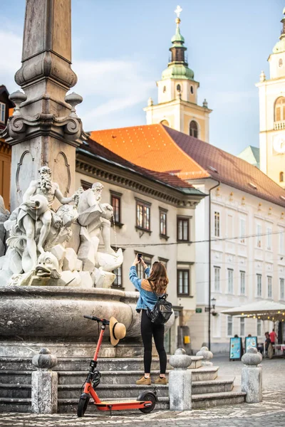 Viajante explorando a cidade velha em scooter elétrico ambientalmente amigável. turista feminina explorando Ljubljanas antigo centro histórico medieval da cidade, tirando fotos de uma fonte com seu telefone. — Fotografia de Stock
