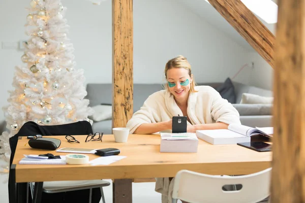 Jonge zakenvrouw met een warme badjas en cosmetische ooglapjes die in de winter in de winter op afstand werken Kerst in 2020 corona virus pandemie. Werk van thuis, selter op zijn plaats, concept — Stockfoto