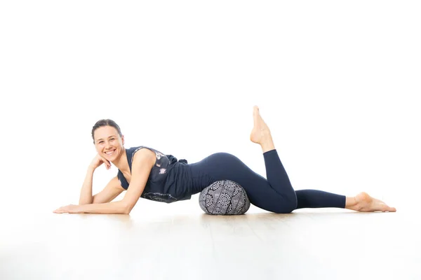 Restorative yoga with a bolster. Young sporty female yoga instructor in bright white yoga studio, lying on bolster cushion, stretching, smilling, showing love and passion for restorative yoga — Stock Photo, Image