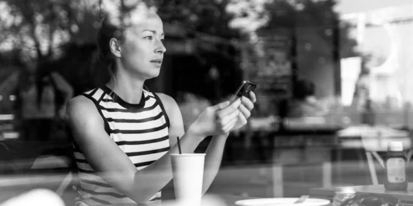 Kahve molasında kahve dükkanının camından bakarken cep telefonu tutan düşünceli beyaz kadın.. — Stok fotoğraf