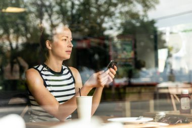 Kahve molasında kahve dükkanının camından bakarken cep telefonu tutan düşünceli beyaz kadın..