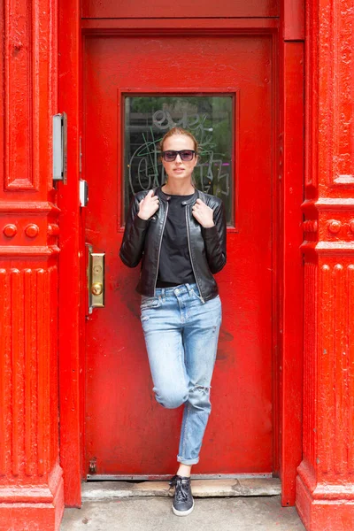 Mujer joven de moda con gafas de sol, chaqueta de cuero negro y pantalones vaqueros Bleu posando frente a la puerta roja en la calle de la ciudad de Nueva York. —  Fotos de Stock