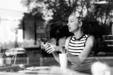 Kahve molasında kahve dükkanının camından bakarken cep telefonu tutan düşünceli beyaz kadın..