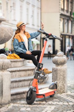 Şehir merkezindeki eski tarihi binanın merdivenlerinde oturan ve telefonuyla fotoğraf çeken genç bir kadın. Elektrikli scooterla Ljubljanas 'ın eski kasabasını keşfeden bir kadın gezgin. Yaz tatili.