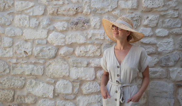 Retrato de una hermosa mujer rubia alegre con un vestido de verano de una pieza y un sombrero de verano, de pie frente a la antigua pared de piedra medieval. concepto de retrato de vacaciones de verano. —  Fotos de Stock