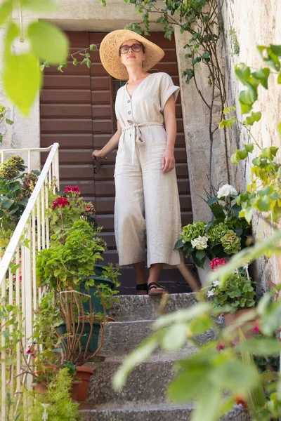 Hermosa turista femenina con sombrero de sol de paja grande y vestido de pie en la vieja escalera de piedra vintage casa durante el viaje de verano en el costo mediterráneo en el día caluroso de verano. —  Fotos de Stock