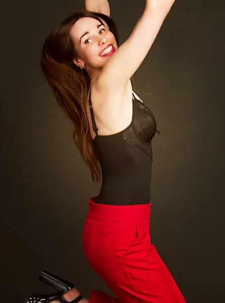 Studio portrait of a young woman in a jump, in red trousers and a black bodysuit, on a dark background. Warm toning.