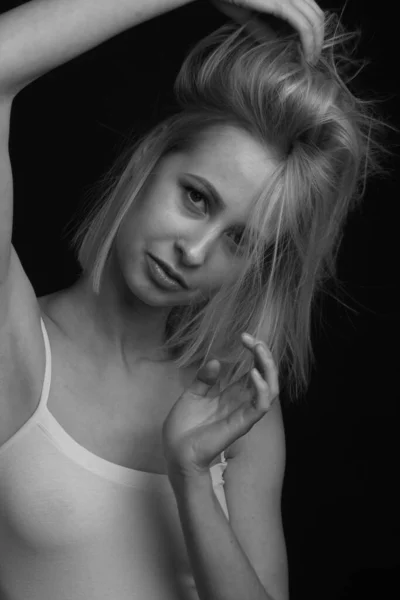 black-and-white portrait of a blonde on a black background. in a white tank top with straps