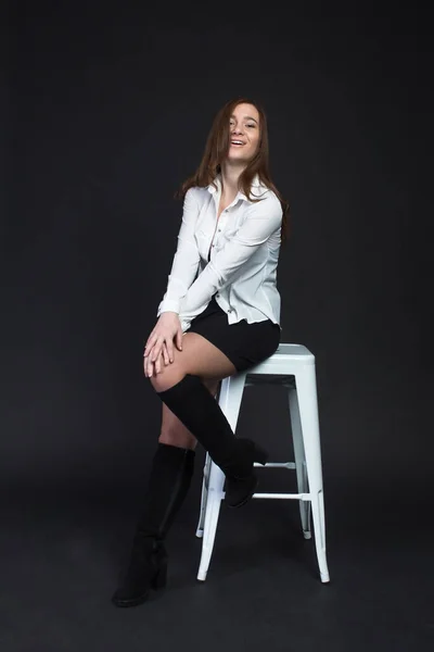 Young brown-haired woman, studio photo on a black background, sitting on a white chair in a white blouse