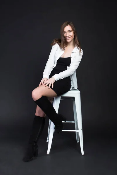 Young brown-haired woman, studio photo on a black background, sitting on a white chair in a white blouse