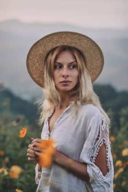 Keten elbiseli sarışın dağlardaki bir çayırda otların ve kır çiçeklerinin arasında yürüyor.