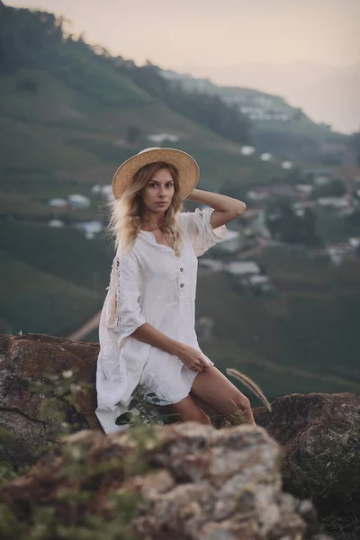 Den blonda i linneklänning och halmhatt sitter på en utsiktsplats i bergen — Stockfoto