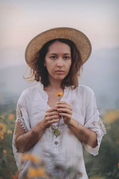 Keten elbiseli ve hasır şapkalı genç bir kadın dağlarda bir bakış açısında oturuyor. — Stok fotoğraf