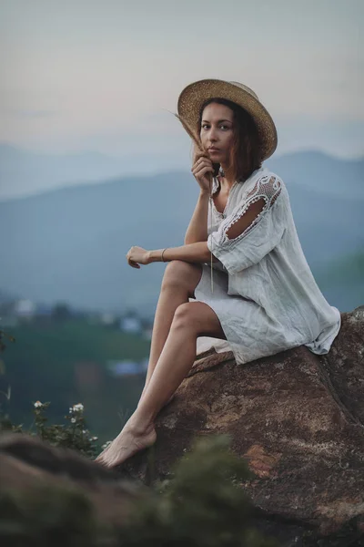 Blondynka w lnianej sukience i słomkowym kapeluszu siedzi na widoku w górach — Zdjęcie stockowe