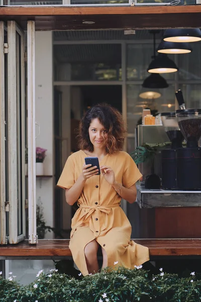 在咖啡店的阳台上，穿着黄色连衣裙的年轻女人用她的智能手机 — 图库照片