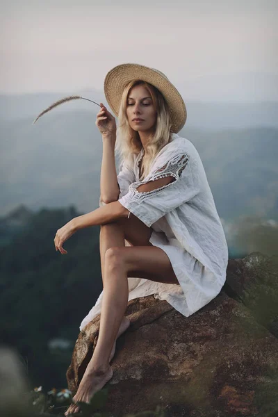 Keten elbiseli ve hasır şapkalı sarışın dağlarda bir bakış açısında oturuyor. — Stok fotoğraf