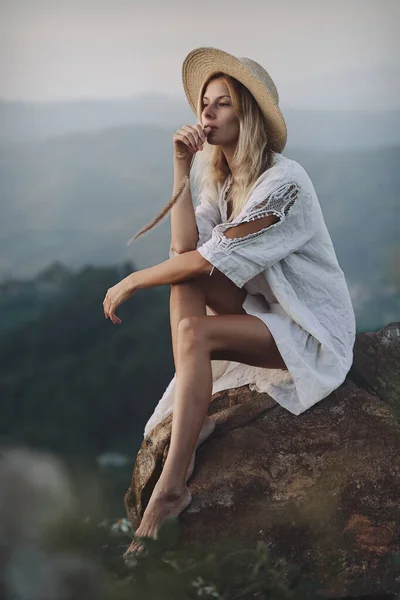 Η ξανθιά με το λινό φόρεμα και το ψάθινο καπέλο κάθεται στα βουνά. — Φωτογραφία Αρχείου