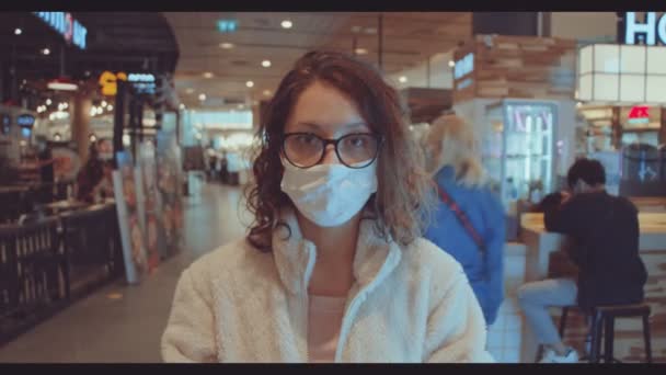 Vrouw met gezichtsmasker en kijkend naar de camera in het winkelcentrum. vrouw in witte trui met gezichtsmasker in winkelcentrum — Stockvideo