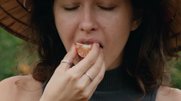 Uma mulher turista fica em um jardim laranja usando um chapéu vietnamita e come uma laranja descascada de uma árvore — Vídeo de Stock