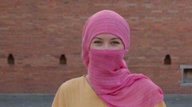 Doğal makyajlı, pembe başörtüsü takan çekici Müslüman kadın kameraya bakıyor.