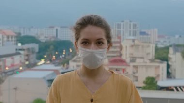 Koruyucu tıbbi maske takan ve çatıda duran güzel bir genç kadının portresi. Bulanık Şehir Arkaplanı