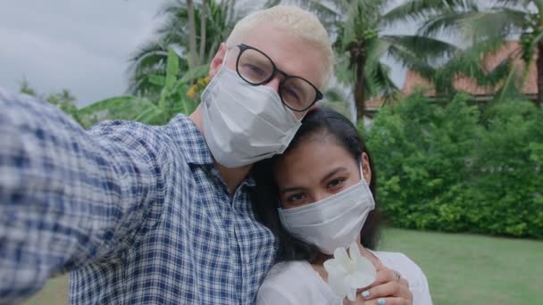 若い国際的なカップルが自撮りをする。安全な旅行は新しい通常です。公園で顔のマスクをしたヨーロッパ人男性とアジア人女性。新しい正常なウイルスの概念を旅行する — ストック動画