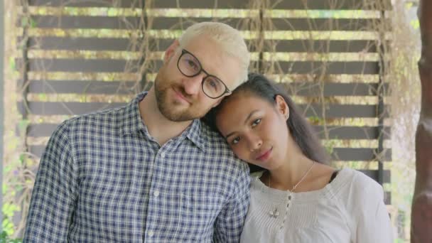 Ένα νεαρό διεθνές διαφυλετικό ζευγάρι ερωτευμένο κοιτάζει κατευθείαν στην κάμερα μια ηλιόλουστη μέρα στον κήπο του σπιτιού. — Αρχείο Βίντεο