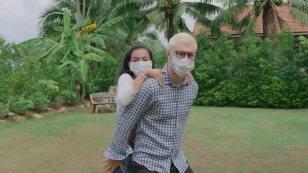 Jovem casal misto vestindo máscara protetora correndo piggyback e se divertindo no jardim de casa — Vídeo de Stock