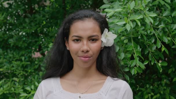 Портрет молодой красивой тайской женщины с цветком в волосах, стоящей в саду и смотрящей прямо в камеру в солнечный день — стоковое видео