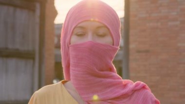 Doğal makyajlı, pembe başörtüsü takan çekici Müslüman kadın kameraya bakıyor.