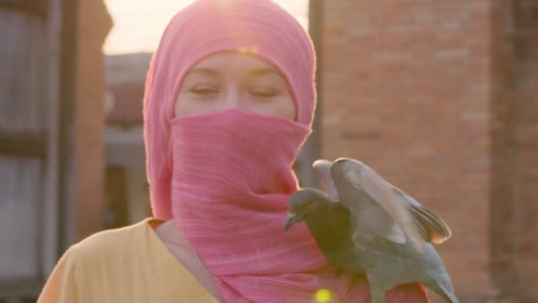 Приваблива мусульманка з натуральним гримом у рожевому шарфі хіджабу стоїть з сонцем на задньому плані і дивиться на камеру, в той час як голуб сидів на плечі дівчаток. — стокове відео