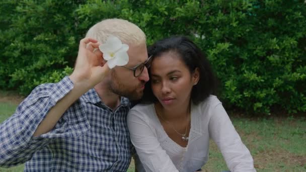 Jeune couple international amoureux assis sur l'herbe et un homme fumant une fleur comme une blague par une journée ensoleillée dans le jardin de la maison — Video