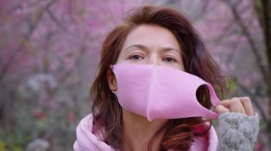 Pembe kapüşonlu ve pembe koruyucu maskeli bir kadın Sakura bahçesinde duruyor ve doğrudan kameraya bakıyor.