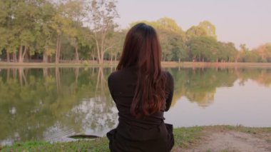 Taylandlı genç kadın gün batımında gölün kenarında dalgın dalgın oturuyor.