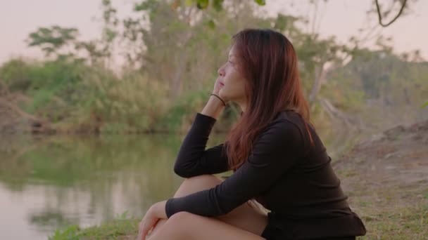 Thaise jonge vrouw zit gepensioneerd door meer in park bij zonsondergang — Stockvideo