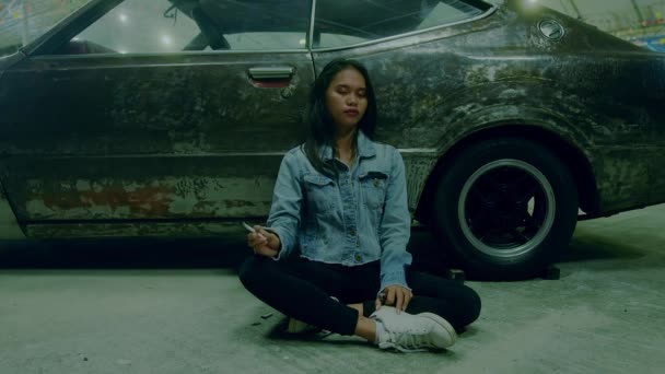 Ein trauriges thailändisches Mädchen sitzt rauchend an ein rostiges Auto gelehnt — Stockvideo
