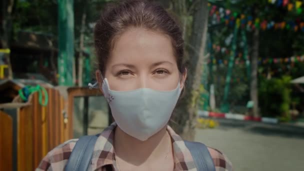 Mujer joven en la máscara médica de moda protectora en su cara mirando a la cámara al aire libre — Vídeo de stock