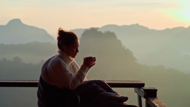 Joven mujer está sentada con una taza de café en un acantilado con vistas a las montañas con niebla en la mañana — Vídeos de Stock