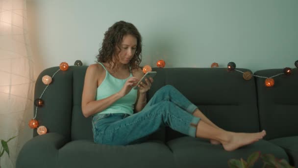 Jeune femme utilise un téléphone intelligent et s'assoit sur le canapé tranquillement dans le salon — Video