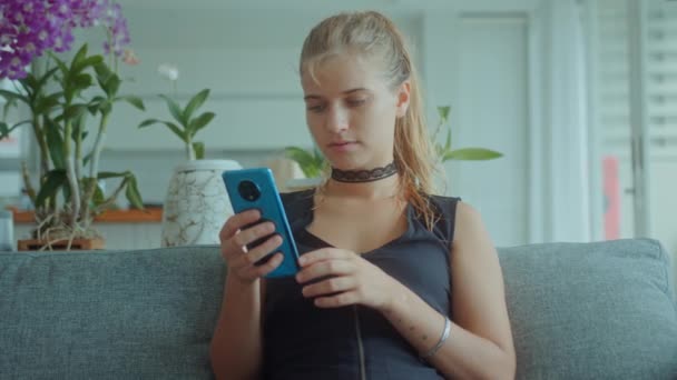 Wunderschöne junge blonde Frau sitzt auf einem Sofa und trägt ein sexy schwarzes Kleid mit Choker am Hals, während sie mit dem Handy Dating-Apps anschaut und verführerische Nachrichten verschickt — Stockvideo