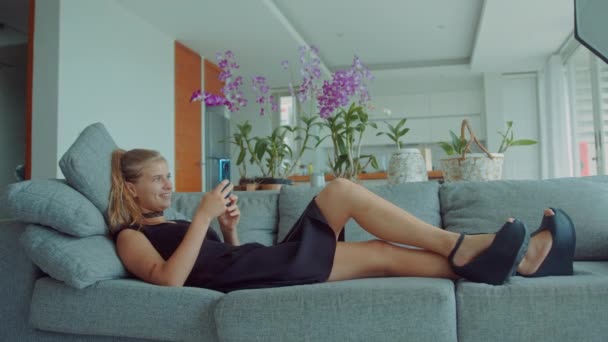 Magnifique jeune femme blonde allongée sur un canapé portant une robe noire sexy avec étranglement sur son cou et en utilisant un téléphone mobile en regardant sortir ensemble applications comme elle glisse et envoie des messages séduisants — Video