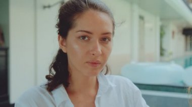 Kahverengi saçlı ve güzel kahverengi gözlü genç bir kadın otelin avlusunda oturmuş kameraya gülümsüyor.