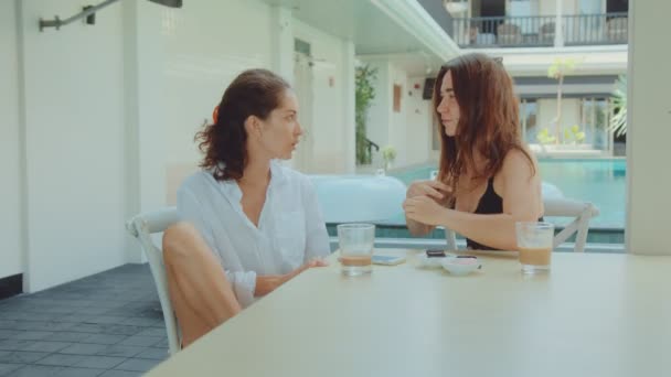 Due giovani donne che parlano a un tavolo in un hotel con una piscina sullo sfondo — Video Stock