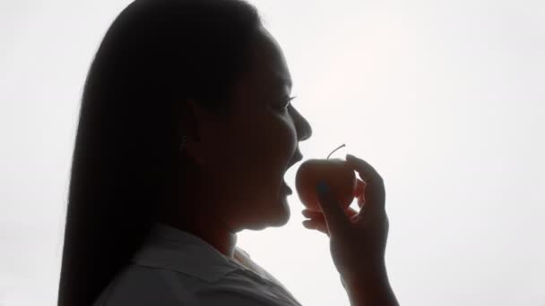 En silhuett av en ansenlig kvinna i vit skjorta tar en bit av ett äpple och tittar sedan på kameran — Stockvideo