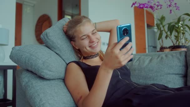 Nádherné blondýny žena ležící na pohovce na sobě sexy černé šaty s choker na krku a pomocí mobilního telefonu při pohledu na randění aplikací, jak ona swipes a posílá svůdné vzkazy — Stock video