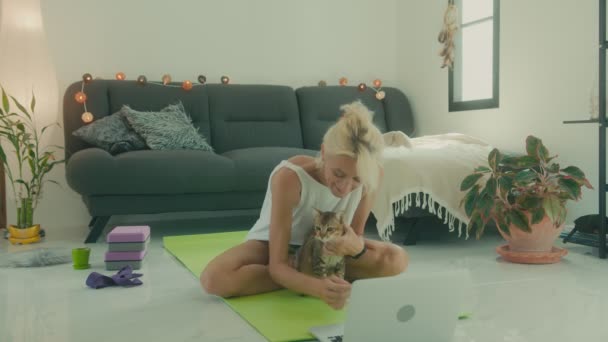 Dojrzała kobieta siada z kotem na macie do jogi przed laptopem w ramach przygotowań do zajęć jogi online. — Wideo stockowe