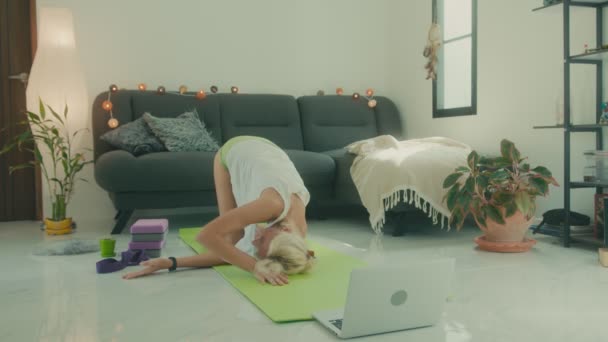 Mujer haciendo estiramiento en el suelo en casa delante de su portátil, clase de yoga en línea — Vídeos de Stock